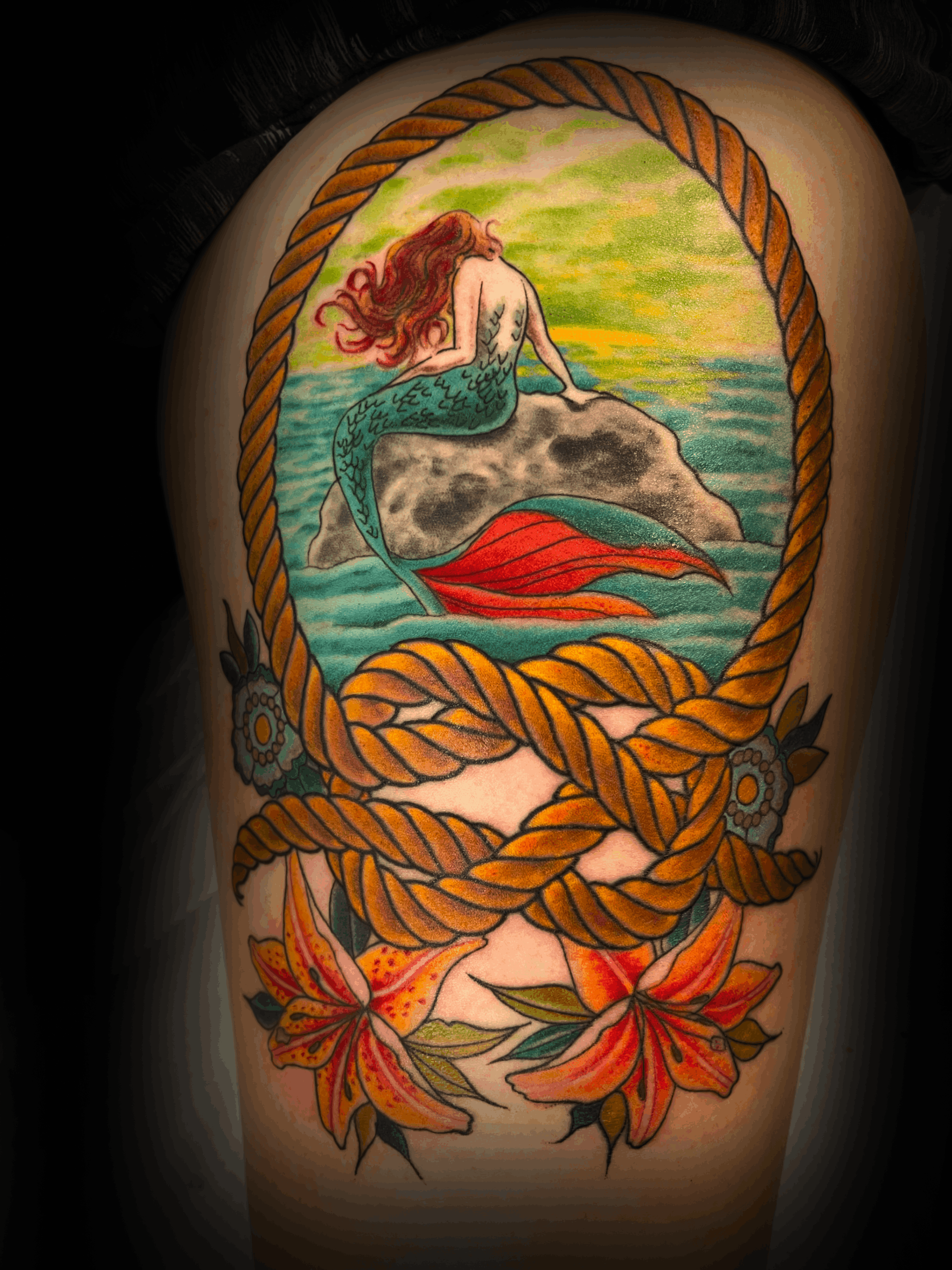 An image of a custom tattoo from Kevin Younger's Lucky Cat Tattoo Parlour: a design featuring a mermaid sitting on a rock within a rope frame
