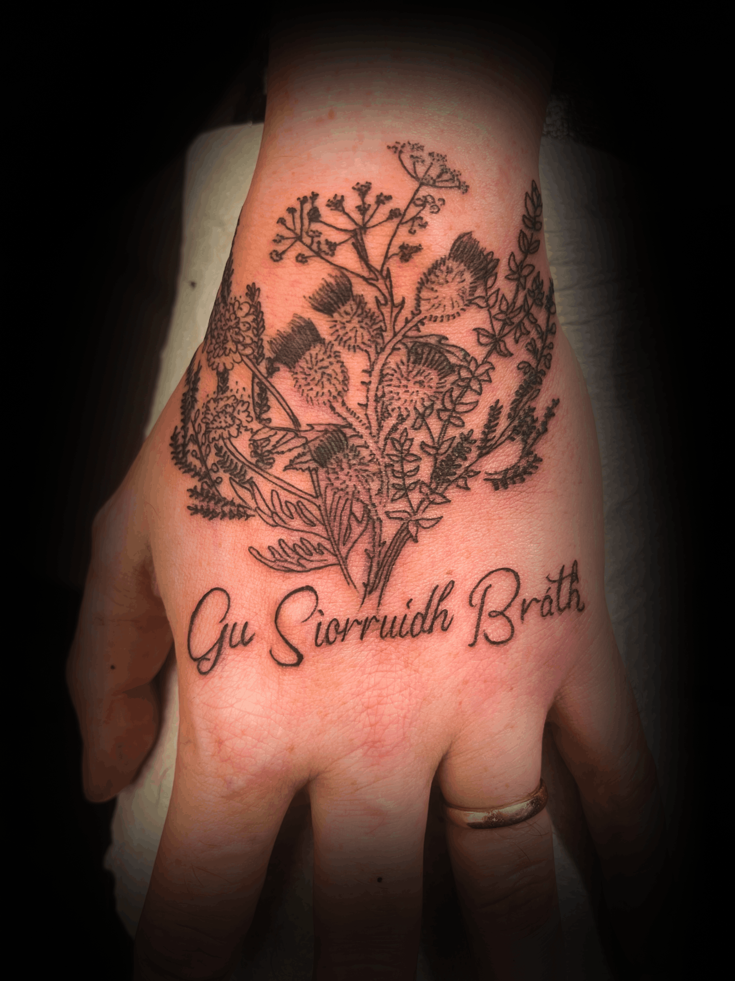 An image of a custom tattoo from Kevin Younger's Lucky Cat Tattoo Parlour: a design featuring a black and grey Thistles on a hand with script text below